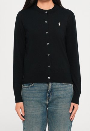 Femme portant un cardigan noir cintré boutonné à manches longues et un jean bleu, debout devant un fond blanc uni.