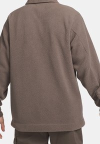 Hnědá fleecová bunda s texturovaným povrchem, dlouhými rukávy, límcem a volným střihem, navržená pro teplo a pohodlí.