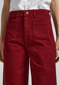 Punaiset puuvillaiset leveälahkeiset housut, joissa on nappisuljenta, kaksi suurta takataskua ja sileä, hieman teksturoitu pinta.