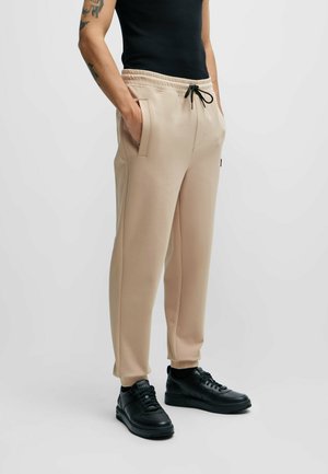 Pantalon de jogging beige avec taille élastique et cordons noirs, doté de poches latérales et d'un design de jambe fuselée, associé à des baskets noires.