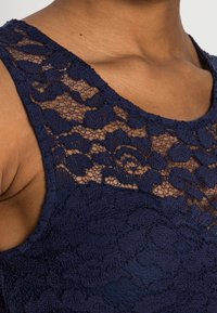 Marinblå spetsblus med rund halsringning, prydd med ett blommönster och texturerat tyg. Ärmlös design med en slät, figurnära bodice.