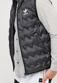 Chaleco de plumas acolchado negro con cierre de cremallera en la parte frontal, que presenta un logo de Adidas en el pecho y una textura suave y brillante.