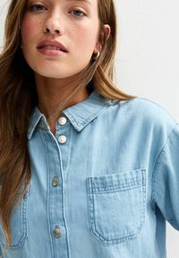 Camisa de mezclilla azul claro con botones en la parte delantera, cuello puntiagudo y un solo bolsillo en el pecho. Presenta botones de presión y una tela ligeramente texturizada.