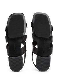 Sandales noires avec une tige en cuir lisse, présentant un design minimaliste et deux brides ajustables. Semelle extérieure en caoutchouc texturé, talon plat.