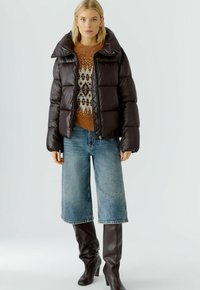 Schwarze Pufferjacke mit hohem Kragen und gestepptem Design, getragen über einem gemusterten braunen Pullover, kombiniert mit hellblauen weit geschnittenen Jeans und braunen knielangen Stiefeln.
