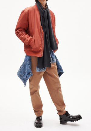 Osoba ubrana w rdzawe bomberkę, czarny szalik, beżowe spodnie, czarne błyszczące buty oraz niebieską kurtkę dżinsową zawiązaną wokół talii.