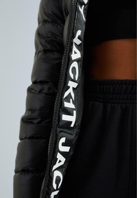 Černá puffa bunda s lesklým povrchem, nabízející zipové zapínání a výrazné bílé logo na vnitřní straně, spárovaná s černými kalhotami.