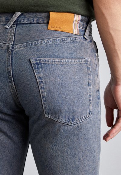 Jeans de mezclilla azul claro con un acabado texturizado, que presentan un bolsillo trasero y una etiqueta de cuero naranja con rayas y el nombre "Paul Smith" en relieve.