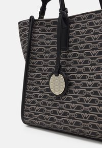 Tote-väska i mönstrad tyg med en svart och beige logotypdesign. Har läderdetaljer och en rund metallisk logotag.