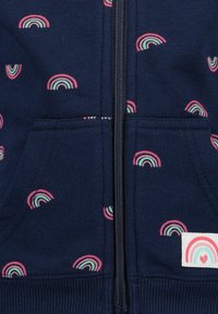 Navy sweatshirt with a front zipper, featuring small, multicolored rainbow patterns and a white tag with a rainbow design and heart.