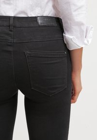 Vaqueros negros de denim suave, con un diseño ajustado, bolsillos traseros y un ligero brillo. Etiqueta de cuero en la cintura con el nombre de la marca.