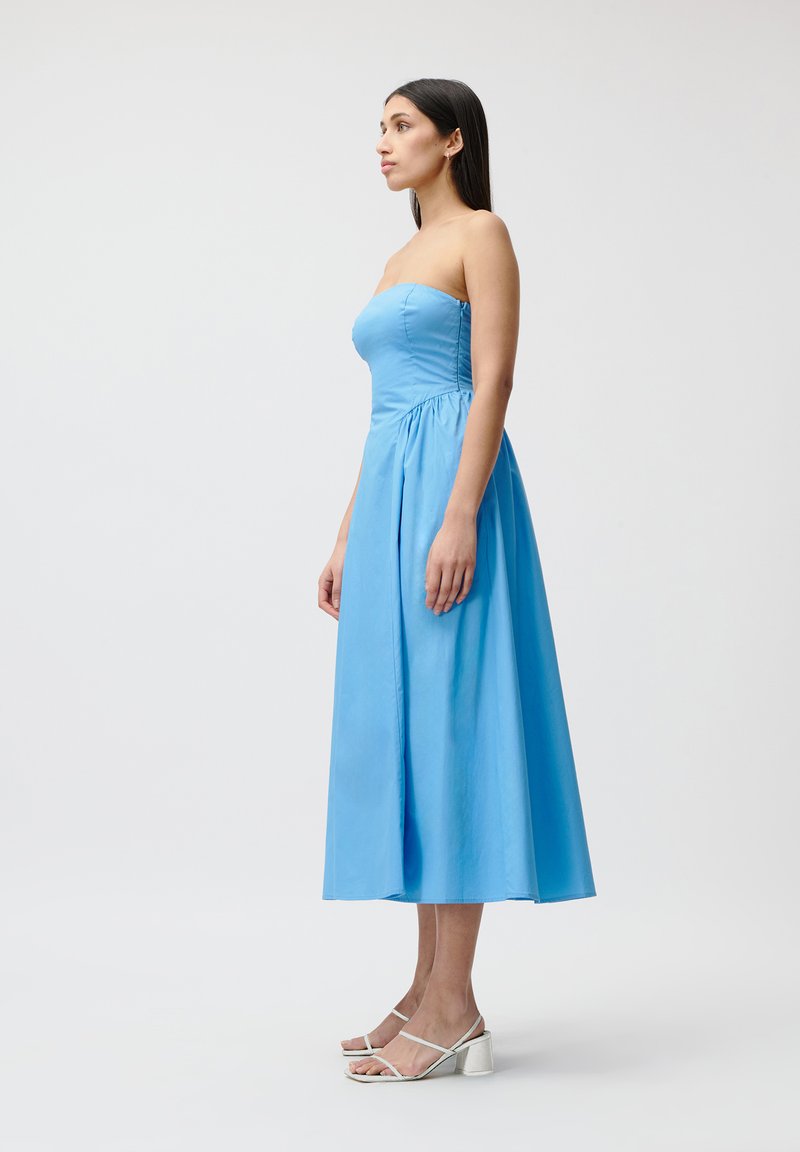 Bezramieniowa niebieska sukienka z lekkiego materiału, z dopasowanym gorsetem i zwiewnym dołem. Posiada marszczoną talię i długość do połowy łydki. W połączeniu z białymi sandałami na słupku.