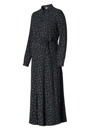Femme portant une robe noire à manches longues avec un imprimé floral vert, une ceinture à la taille, et des bottes noires montant jusqu'aux genoux, debout devant un fond uni.