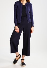 Tmavě modrý kardigan s knoflíky, černý přiléhavý top, tmavě modré široké kalhoty a černé dřeváky se zapínáním kolem kotníku a světlou podrážkou.