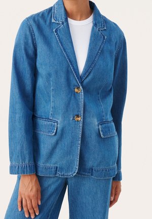 Personne portant un blazer en denim bleu moyen avec deux boutons et des poches à rabat, associé à un pantalon en denim assorti et une chemise blanche en dessous.