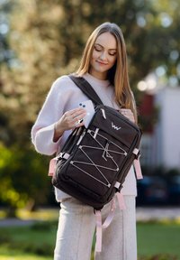 Černý batoh s růžovými doplňky a detailem z pružného šňůrky. Má několik přihrádek a drží ho osoba, která si bere telefon.