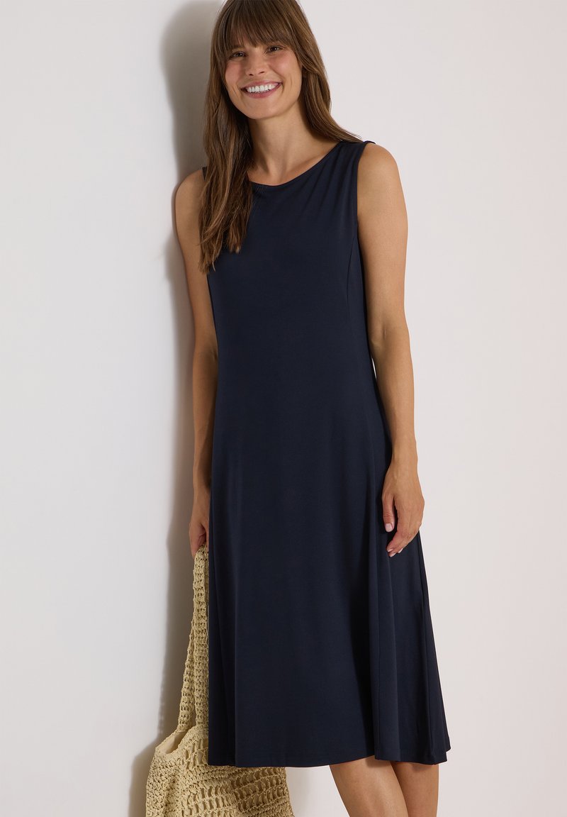 Femme aux longs cheveux bruns portant une robe midi bleu marine sans manches, tenant un sac fourre-tout tissé beige, debout et souriante devant un mur blanc.