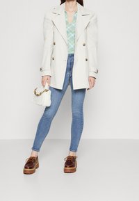 Abrigo trench de crema con botones dorados sobre una blusa de cuadro azul-verde. Jeans ajustados azules y zapatos de plataforma marrones. Bolso blanco texturizado con cadena.