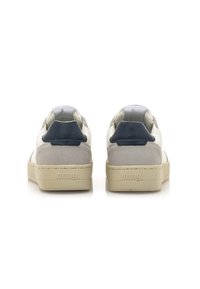 Las zapatillas presentan una combinación de cuero blanco y gamuza gris claro, con acentos en azul marino en el talón y un interior suave, montadas sobre una suela beige.