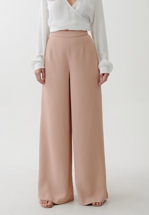 Pantaloni a gamba larga in tessuto rosa chiaro, a vita alta con una texture morbida, abbinati a un top bianco a maniche lunghe. Nessun sistema di chiusura visibile.