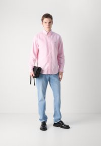 Lyserød knappet skjorte, lys blå jeans, sorte sko, holder en sort veske. Enkelt design, glatt tekstur, ingen synlige mønstre.