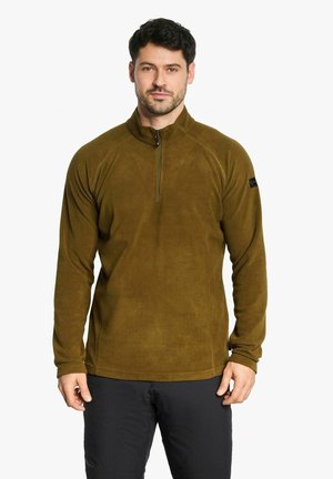 Homme portant un pull polaire marron avec fermeture éclair quart et un pantalon noir, debout sur un fond blanc uni.