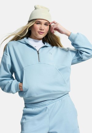 Jonge vrouw die een lichtblauw hoodie- en joggingpakken draagt met een witte muts, poseert met één hand in haar zak en de andere vlak bij haar gezicht.