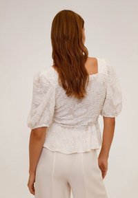 Blusa bianca strutturata con maniche a palloncino e motivo floreale. Presenta uno scollo quadrato sulla schiena e una vita a peplo, abbinata a pantaloni chiari.