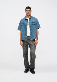 Blå denimskjorta med kort ärm och två bröstfickor, i kombination med grå vida byxor och svarta skor. Enkel grafisk t-shirt under.