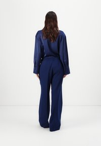 Camicia lunga in satin blu navy con vestibilità oversize, abbinata a pantaloni a gamba larga a vita alta in tessuto blu navy coordinato. Texture liscia, design minimal.