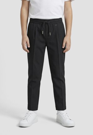 Persoon draagt zwarte taps toelopende broek met koord in de taille en witte sneakers, staand voor een effen lichte achtergrond.