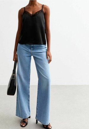 Personne portant un top caraco noir bordé de dentelle, un jean large bleu clair, des talons ouverts noirs et tenant un sac à main noir, debout sur un sol uni.