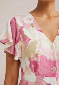 Květinové šaty s velkými růžovými a zlatými květinovými vzory na bílém pozadí, V-výstřihem, krátkými volánkovými rukávy a knoflíkovými detaily.