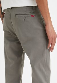 Man med grå Levi's byxor som visar bakfickan med knapp och varumärkesetiketter, vit skjorta och tatuerad arm synlig.