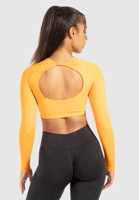 Langärmliges cropped Top in leuchtendem Orange mit einem runden Ausschnitt auf der Rückseite. Glatte Textur, taillierte Passform, kombiniert mit hoch taillierten schwarzen Leggings.