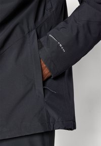 Μαύρο αδιάβροχο μπουφάν με ψηλό γιακά, κλείσιμο καρπού με βέλκρο και λογότυπο OMNI-TECH στο μανίκι. Υφασμάτινη υφή και πλευρικές τσέπες.