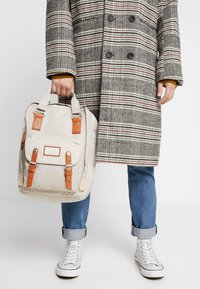 Sac à dos beige avec des accents en cuir brun, doté d'une poche avant, de quincaillerie en argent et de deux poignées supérieures. Porté avec un manteau à carreaux et un jean.