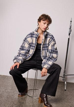 Personne aux cheveux courts portant une veste polaire à carreaux, un pantalon noir et des bottes marron, assise sur un tabouret en métal devant un fond uni.