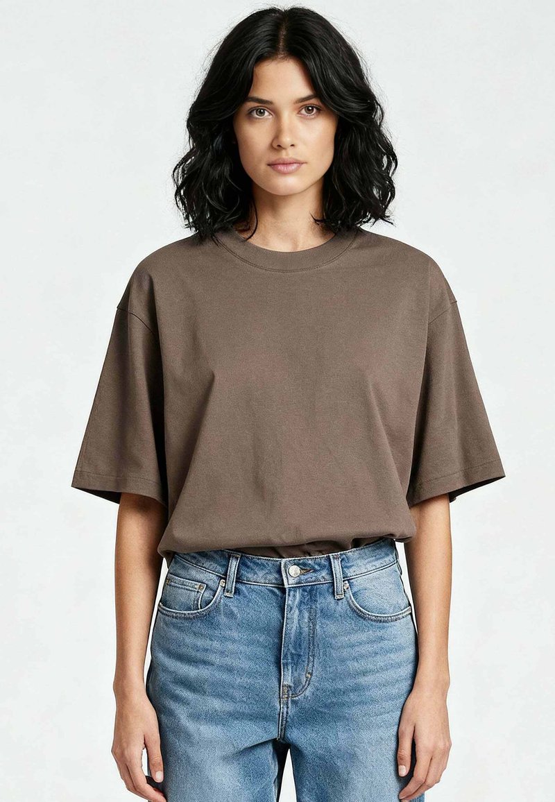 Femme aux cheveux noirs mi-longs portant un T-shirt marron ample rentré dans un jean bleu taille haute, debout devant un fond uni.