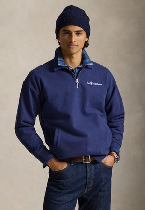 Mladý muž v tmavomodrej čiapke, tmavomodrej polozipovej mikine cez modrú károvanú košeľu, tmavých džínsoch, hnedom opasku a náramkových hodinkách, pózujúci s rukami v vreckách.