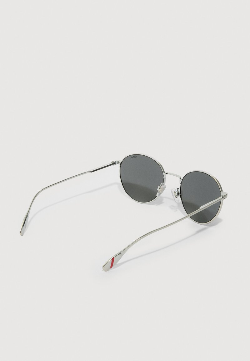 Silver metal sunglasses with round black lenses. Slim arms feature a red accent. Design emphasizes minimalism with a sleek silhouette.