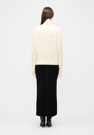 Γυναίκα με μακριά καστανά μαλλιά, που φοράει κρεμ πουλόβερ με ζιβάγκο, μαύρη μακριά φούστα και μαύρα μπότες με τακούνι, στέκεται με την πλάτη προς τα εμπρός σε λευκό φόντο.