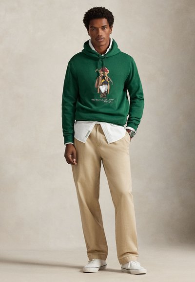 Zelena majica s kapuljačom s grafičkim printom medvjeda, preko bijele košulje s ovratnikom, uparena s bež hlačama i bijelim tenisicama.