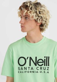 Mėtų žalios spalvos trumpomis rankovėmis marškinėliai, kurių priekyje užrašyta ryškiais juodaisiais rašmenimis "O'Neill SANTA CRUZ CALIFORNIA-U.S.A.". Pagaminti iš medvilnės.