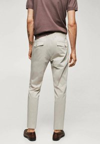 Pantalons slim beige avec deux poches arrière à rabat, accents de bouton et une texture lisse, associés à des chaussures brunes.
