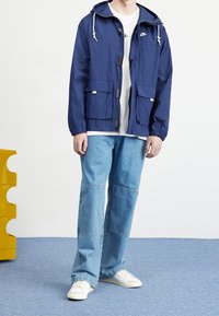 Giacca leggera della Marina con cappuccio, zip anteriore e due grandi tasche. Abbinata a jeans in denim a gamba larga e scarpe da ginnastica bianche su uno sfondo blu.