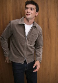 Brown corduroy jacket with a zipper and a chest pocket, worn over a white T-shirt, paired with dark denim jeans against a wood background.