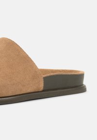 Καφέ σουέντ παπούτσι κλειστού τύπου, χωρίς κορδόνια, με επίπεδη μαύρη σόλα και χαμηλό τακούνι σε λευκό φόντο.