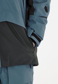 Blau-schwarze, wetterfeste Jacke mit strukturierter Oberfläche, ausgestattet mit einer Reißverschlusstasche und Klettakzenten an den Manschetten.