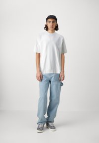 Weißes T-Shirt mit kurzen Ärmeln kombiniert mit hellblauen Jeans und grauen Sneakers. Dazu kommt eine schwarze Kappe, vor einem einfachen weißen Hintergrund.